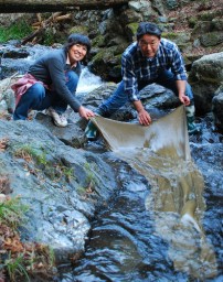 妻、ひろ子さん(左)と蚕のふんで染めた布を山田川に広げる松井さん